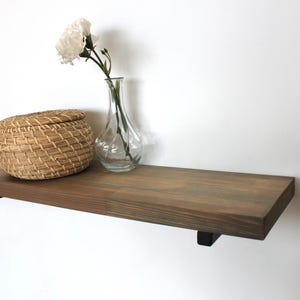 May include: A wooden floating shelf with black brackets, featuring a woven basket and a clear glass vase with white flowers. The shelf is stained in shades of brown and grey.