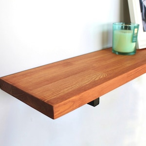 May include: A wooden floating shelf with a warm, reddish-brown finish. The shelf is mounted on a white wall and supported by a black bracket. A framed photo and a green candle in a glass holder sit on the shelf. The shelf is approximately 76 cm long.