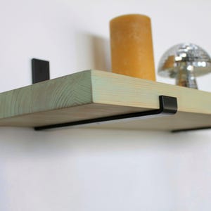 May include: A wooden floating shelf with a pale green edge, held by black metal brackets. An orange candle and a silver disco ball ornament sit on the shelf. The shelf is mounted on a white wall.