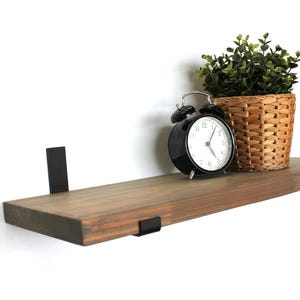 May include: A wooden floating shelf with black metal brackets, holding a black alarm clock, a woven basket, and a small green plant. The shelf is stained a warm brown colour and is mounted on a white wall.