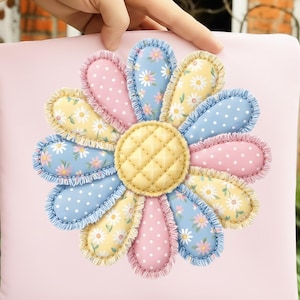 May include: A cushion featuring a large daisy applique. The flower has petals in pink, yellow, and blue with polka dots and floral patterns. The centre is quilted yellow, and the cushion has a light pink background.