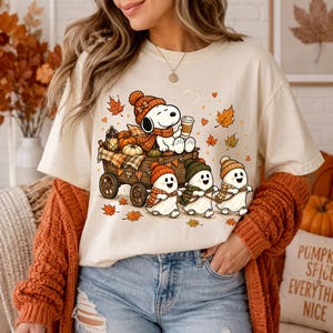May include: A cream-colored t-shirt featuring a graphic of Snoopy sitting in a wooden wagon filled with pumpkins and a lantern, while three ghosts wearing knit hats and scarves pull the wagon, surrounded by falling autumn leaves and hearts.