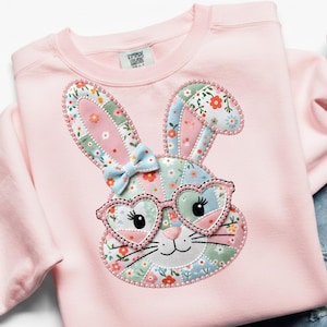 Puede incluir: Sudadera rosa claro con un diseño de conejo de retazos. El conejo tiene gafas en forma de corazón y un lazo azul. Las orejas y la cara presentan estampados florales en rosa, azul y verde. Cuello redondo y mangas largas.
