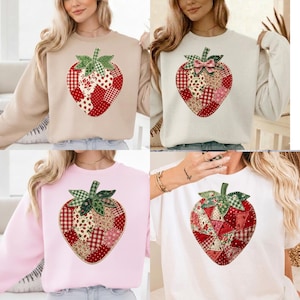 Puede incluir: Una colección de sudaderas y una camiseta, cada una con un gran diseño de fresa. Las fresas están hechas de tela de patchwork en tonos rojos, rosas y beige, con hojas verdes. Las camisetas son en beige, blanco, rosa y blanco.