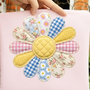 Könnte beinhalten: Ein hellrosa Kissen mit einem großen, applizierten Gänseblümchen-Design. Das Gänseblümchen hat Blütenblätter in Gelb, blauem Vichy-Karo, rosa Vichy-Karo und floralen Mustern. Die Mitte ist gelb gesteppt. Das Design ist handgenäht mit weißem Faden.
