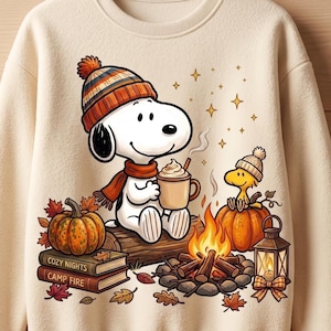 May include: A cream-colored sweatshirt featuring a graphic of Snoopy wearing a striped beanie and scarf, holding a mug of cocoa. Beside Snoopy are pumpkins, a campfire, a lantern, and two books labeled "COZY NIGHTS" and "CAMP FIRE" with autumn leaves.