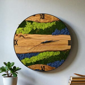 May include: A round wall clock with a natural wood face, featuring green and blue moss accents. The clock has black Roman numerals and hands. The clock is mounted on a gray wall above a wooden shelf with a potted plant and books.