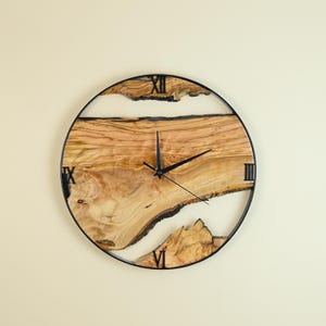 Può includere: Un orologio da parete rotondo con cornice in metallo nero. Il quadrante presenta un design a venatura del legno naturale con accenti di resina bianca. I numeri romani segnano le ore. Le lancette dell'orologio sono nere.