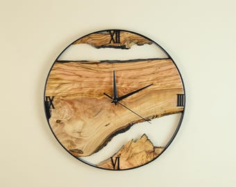 May include: A round wall clock with a black metal frame. The clock face features a natural wood grain design with white resin accents. Roman numerals mark the hours. The clock hands are black.