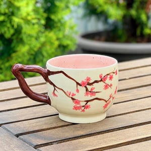 May include: Cream-colored ceramic mug with a brown handle and a pink interior. The mug is decorated with a branch design and pink cherry blossoms. The mug is sitting on a wooden table.