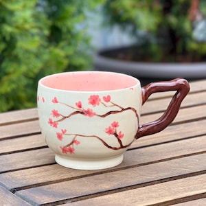 Pode incluir: Uma caneca de cerâmica com exterior creme apresentando ramos castanhos pintados à mão e flores de cerejeira cor-de-rosa. O interior da caneca é de um rosa pastel suave e inclui uma pega grossa, brilhante e castanha escura angular.