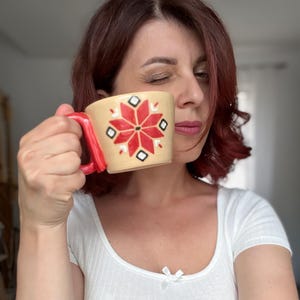 Op de afbeelding: Een beige keramische mok met een rode handgreep en een bloemmotief in rood, zwart en wit. Het bloemmotief is gecentreerd op de voorkant van de mok. De mok wordt in de hand van een persoon gehouden.