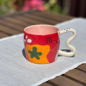 May include: A ceramic mug with a unique, wavy handle. The mug features a vibrant design with red, yellow, and green abstract shapes. White polka dots and black speckles add detail. The interior is pink.