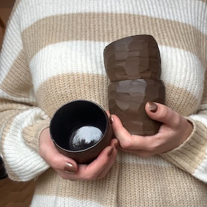 Op de afbeelding: Twee bruine keramische bekers worden vastgehouden voor een beige en wit gestreepte trui. Eén beker is een kleine, ronde kom. De andere is een hogere, getextureerde beker met een gebeeldhouwd uiterlijk. De bekers zijn donkerbruin.