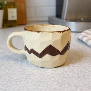 May include: Cream-colored ceramic mug with a geometric design and a brown zigzag pattern. The mug has a rounded handle and a textured surface. The rim and the zigzag pattern are brown. The mug is on a light gray surface.