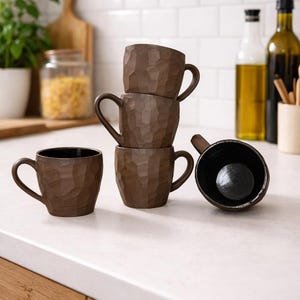 Puede incluir: Cinco tazas de cerámica marrón con exteriores texturizados y asas curvas. Una taza está tumbada de lado. Las tazas están sobre una encimera blanca. En el fondo se ven artículos de cocina.