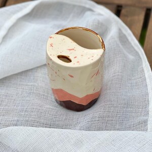 Op de afbeelding: Een keramische tandenborstelhouder met een crèmekleurige basis, besprenkeld met roze verf. De houder heeft een yin-yang ontwerp en is geaccentueerd met roze en bruine lagen. De bovenste rand heeft een bruin glazuur.