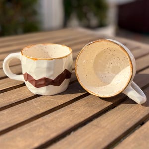 May include: Two handmade ceramic mugs. One upright, the other tilted on its side. Both mugs are white with a geometric brown design and a speckled interior. The rims have a brown trim. The mugs are on a wooden surface.
