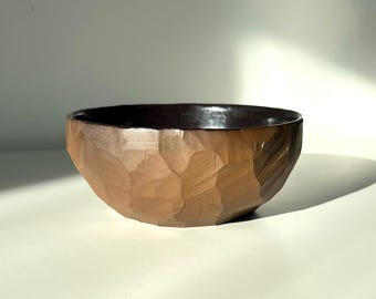 May include: A brown ceramic bowl with a textured exterior and a dark interior. The bowl has a wide, open design, suitable for serving or display. The exterior has a rough, carved appearance.