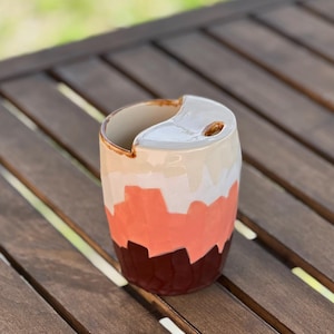 Pode incluir: Uma tigela de cerâmica para fios com um design único. A tigela apresenta um padrão de blocos de cores em tons de castanho, coral, branco e bege. A parte superior tem uma abertura curva e um orifício para o fio. A tigela está numa superfície de madeira.