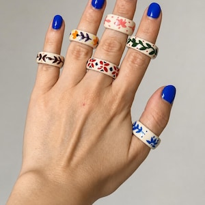 May include: A set of six off-white ceramic rings worn on fingers, each featuring distinct hand-painted patterns including orange flowers, red geometric shapes, green leaves, brown vines, and blue floral motifs in a smooth, glossy finish.