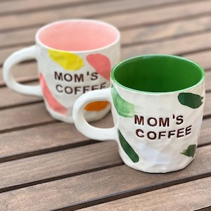 Puede incluir: Dos tazas de café de cerámica con exteriores blancos y asas blancas. Una taza tiene el interior rosa y la otra verde. Ambas tazas tienen el texto "MOM'S COFFEE" y pinceladas coloridas.