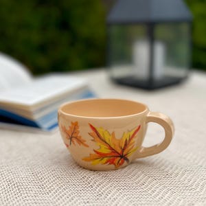 Pode incluir: Caneca de café de cerâmica bege com alça, decorada com folhas de outono pintadas à mão em tons de amarelo, laranja e vermelho. A caneca está sobre uma superfície texturizada esbranquiçada, com um fundo desfocado.