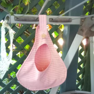 May include: A pink and white striped fabric birdhouse with a circular opening, hanging from a metal structure. The birdhouse has a button detail and is suspended against a green lattice background.