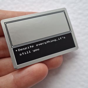 Könnte beinhalten: Ein rechteckiger Metall-Pin mit zweifarbigem Design, bestehend aus einem hellgrauen oberen Bereich und einem schwarzen unteren Bereich. Der schwarze Teil enthält den weißen, verpixelten Text: * Despite everything, it's still you. Silberner Rand.