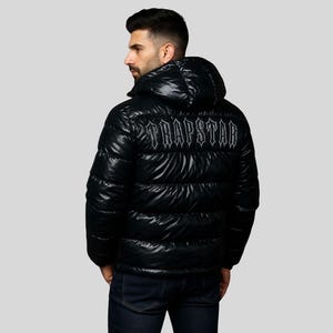 Puede incluir: Chaqueta acolchada negra con capucha. La chaqueta tiene un acabado brillante y presenta la palabra "TRAPSTAR" en letras blancas en la parte superior de la espalda. El modelo lleva vaqueros azul oscuro.