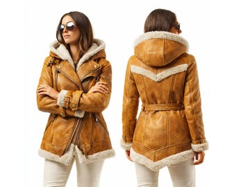 Puede incluir: Chaqueta de cuero marrón con capucha y forro de piel sintética color crema. La chaqueta tiene cierre de cremallera, múltiples bolsillos y cinturón en la cintura. La modelo lleva gafas de sol y pantalones blancos.