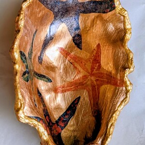 May include: A handmade decorative bowl with a gold exterior and interior. The bowl has a textured surface and is decorated with starfish designs in red, blue, and green. The rim is edged with gold.