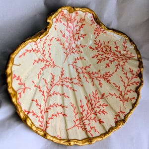 May include: A decorative seashell-shaped dish with a cream interior and a gold-painted rim. The interior features a red coral-like pattern. The dish is likely used for holding small items, such as jewellery or trinkets.