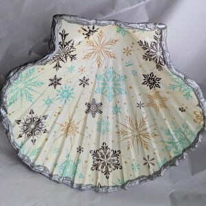 May include: A decorative seashell painted with a snowflake pattern in black, gold, and teal on a cream background. The shell's edge is trimmed with silver paint, creating a festive winter theme.