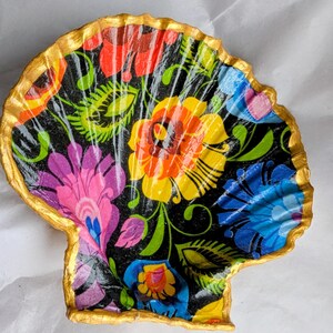 May include: A decorative seashell dish featuring a black background adorned with a colourful floral design in red, orange, yellow, pink, and blue. The rim is edged with gold, enhancing the piece's aesthetic appeal.