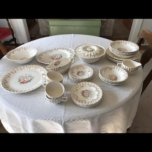 Puede incluir: Un juego de vajilla de porcelana vintage con diseños florales. El juego incluye platos, cuencos, tazas y una fuente para servir con tapa. Los bordes de la vajilla tienen un ribete dorado. La vajilla está dispuesta sobre un mantel blanco.