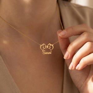 May include: A gold necklace with a pendant shaped like a bow, with the name "Sarah" written in cursive. The necklace is shown against a plain background, emphasizing the intricate design of the jewellery.