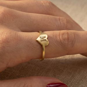 Könnte beinhalten: Ein goldfarbener Ring mit einem herzförmigen Anhänger, auf dem der Buchstabe "M" eingraviert ist. Der Ring wird an einem Finger getragen, wodurch sein elegantes Design hervorgehoben wird. Das Band ist schmal, und das Herz ist der Mittelpunkt.