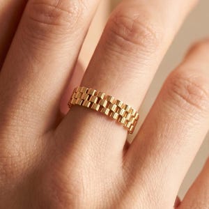 May include: A gold-toned ring featuring a repeating, multi-link chain design that mimics the appearance of a watch bracelet. The band has a uniform width and a polished, metallic finish, creating a textured, segmented look around the finger.