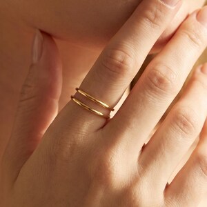 May include: A set of two thin, polished gold rings worn on the middle finger. The rings feature a smooth, rounded band design with a reflective metallic finish. The bands are stacked parallel to each other, creating a simple and uniform appearance.