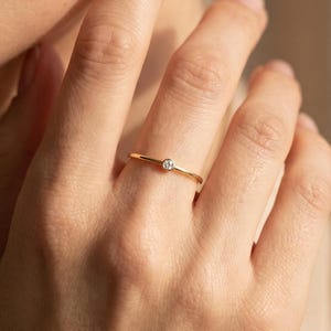 Könnte beinhalten: Ein dünner Goldring mit einem einzelnen, kleinen, runden, klaren Edelstein in einer Zargenfassung. Die Ringschiene ist glatt und poliert und umschließt den Finger mit einem zarten Profil, das das subtile Funkeln des zentralen Steins betont.