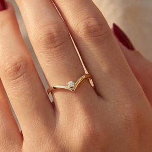 Peut inclure: Une bague dorée en forme de chevron avec une petite pierre précieuse transparente au sommet du V. La bague est portée sur une main avec du vernis à ongles rouge. Le fond est un tissu beige clair.