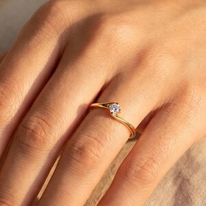 May include: A gold ring with a small, clear gemstone set in a delicate, swirling design. The ring is on a hand with red nail polish, set against a neutral background. The ring's band is thin and elegant.