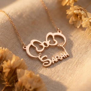 May include: A rose gold necklace with a bow-shaped pendant and the name "Sarah" in a cursive font. The necklace is presented on a beige fabric, with dried flowers adding a decorative touch.