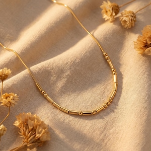 Könnte beinhalten: Eine zarte goldfarbene Halskette mit einer Reihe kleiner, zylindrischer und kugelförmiger Perlen. Die Halskette wird auf einem beigefarbenen Stoff mit getrockneten Blumen im Hintergrund präsentiert. Die Halskette ist ein schlichtes, elegantes Schmuckstück.