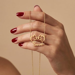 May include: A gold necklace featuring a bow-shaped pendant with the name "SARAH". The pendant and chain are gold-coloured. The necklace is suspended from a fine chain.