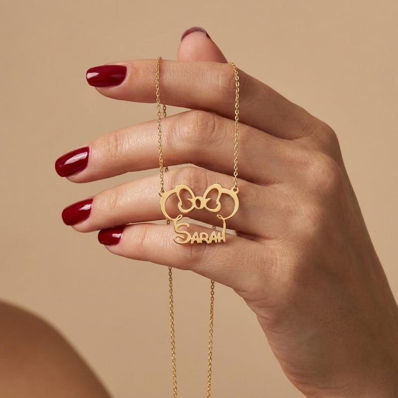 May include: A gold necklace featuring a bow-shaped pendant with the name "SARAH". The pendant and chain are gold-coloured. The necklace is suspended from a fine chain.