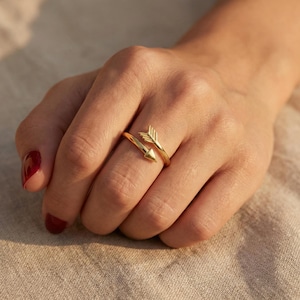Può includere: Un anello a freccia color oro. L'anello presenta un design a freccia, con la punta e le penne della freccia ben visibili. L'anello si avvolge attorno al dito, creando un look unico ed elegante. L'anello è mostrato su una mano.