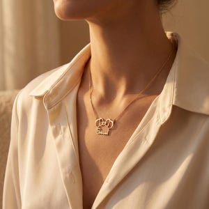 May include: A gold necklace featuring a bow-shaped pendant with the name "Sarah" inscribed below. The necklace is displayed against a cream-coloured blouse.