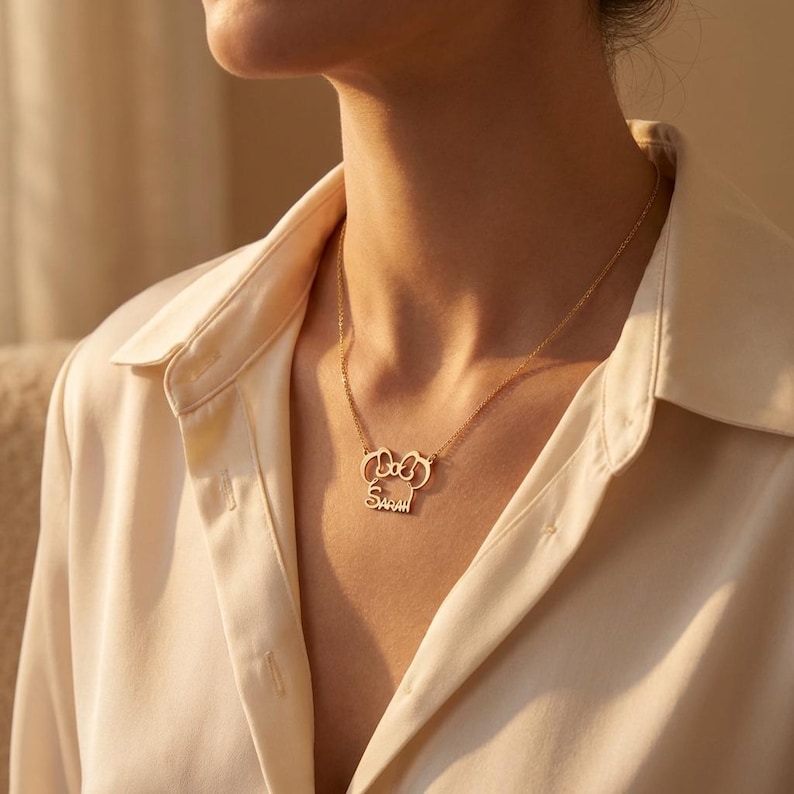 May include: A gold necklace featuring a bow-shaped pendant with the name "Sarah" inscribed below. The necklace is displayed against a cream-coloured blouse.